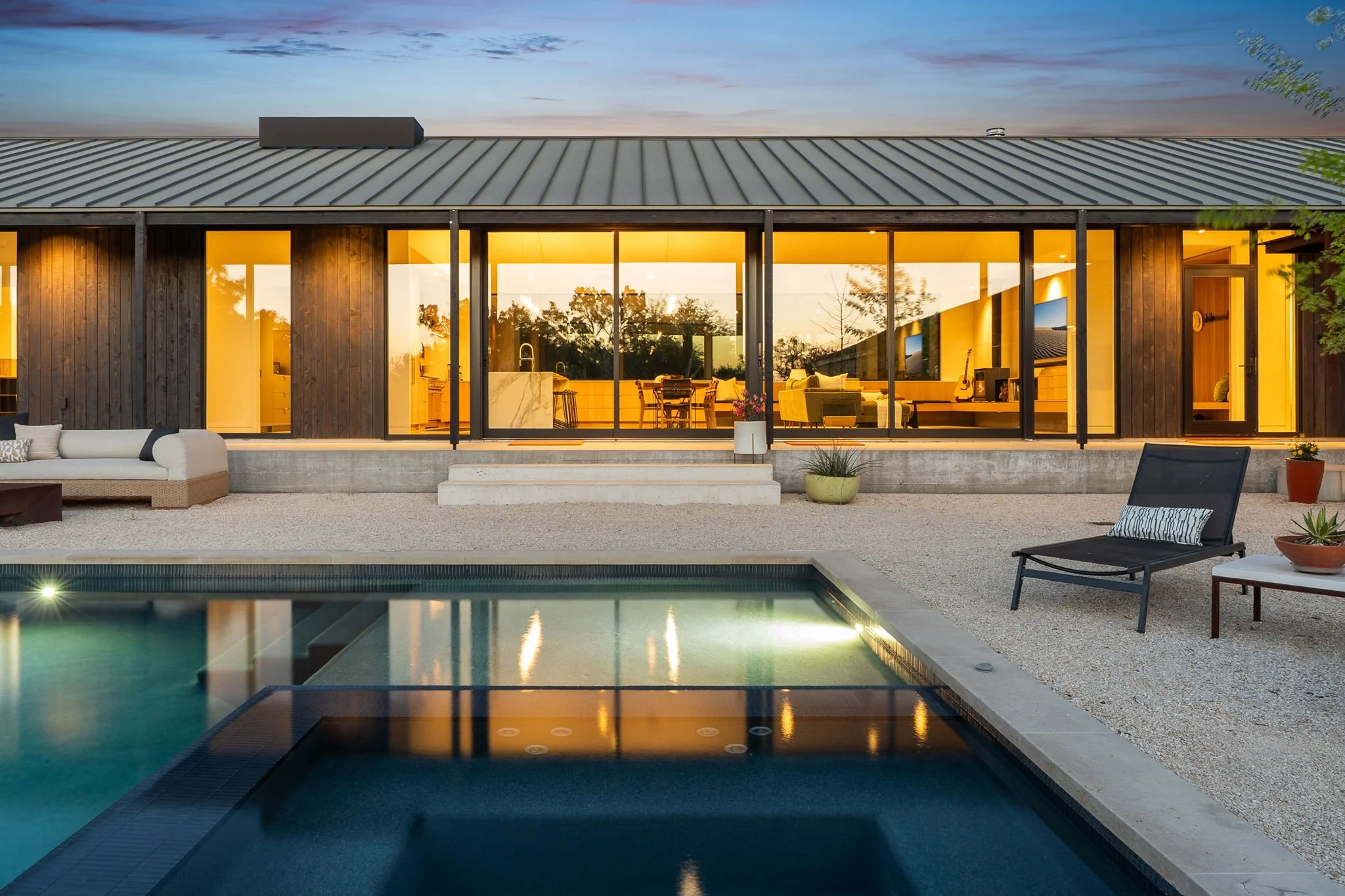 Modern house with large glass windows and interior lighting, a swimming pool in the foreground, outdoor furniture, and trees reflected in the windows during dusk.