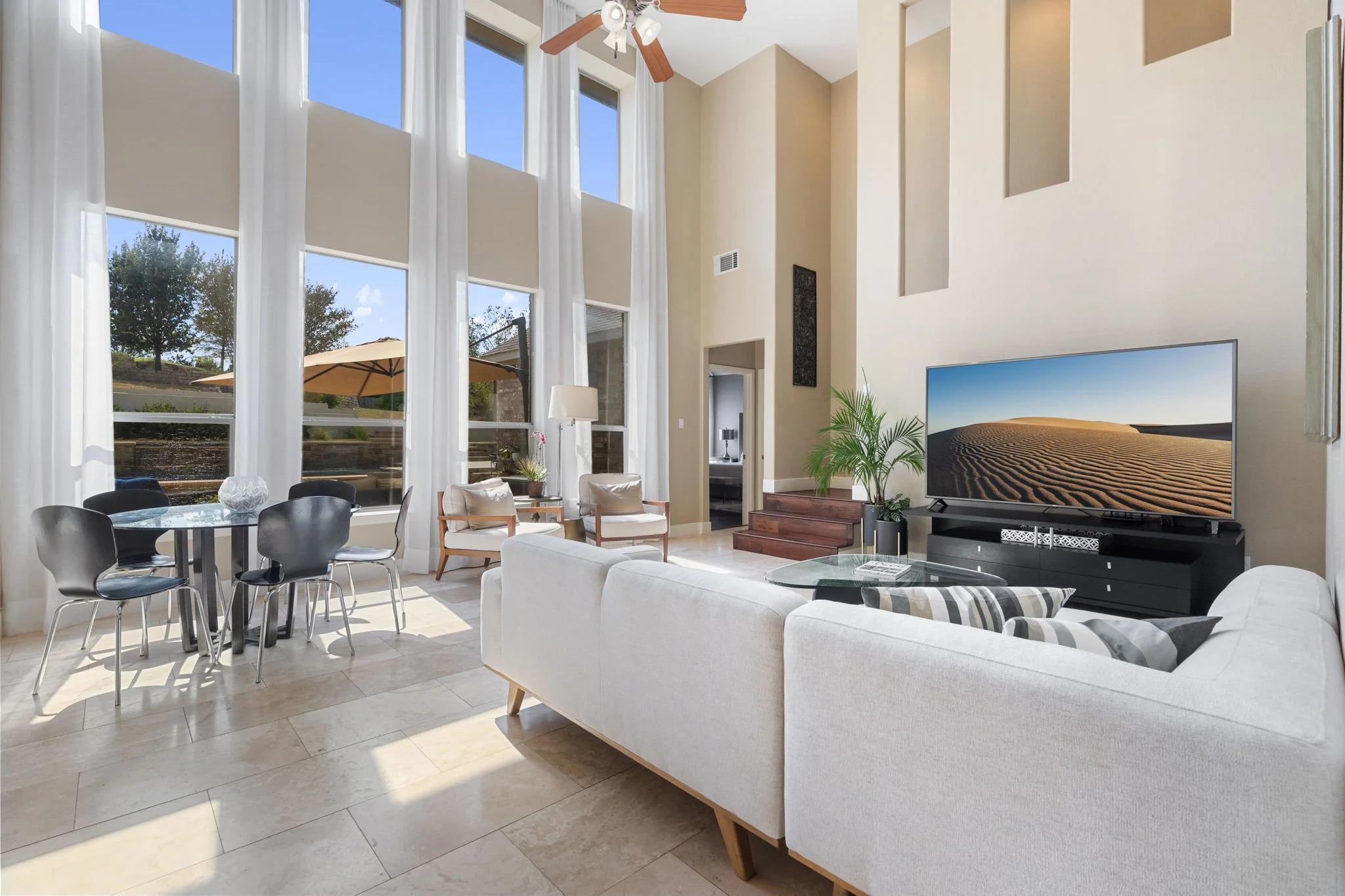 Bright living room with tall ceilings, large windows with white curtains, a ceiling fan, a flat-screen TV on a black stand, white sofa, armchairs, a round dining table with black chairs, indoor plants, and a view of an outdoor patio with a beige umbrella.