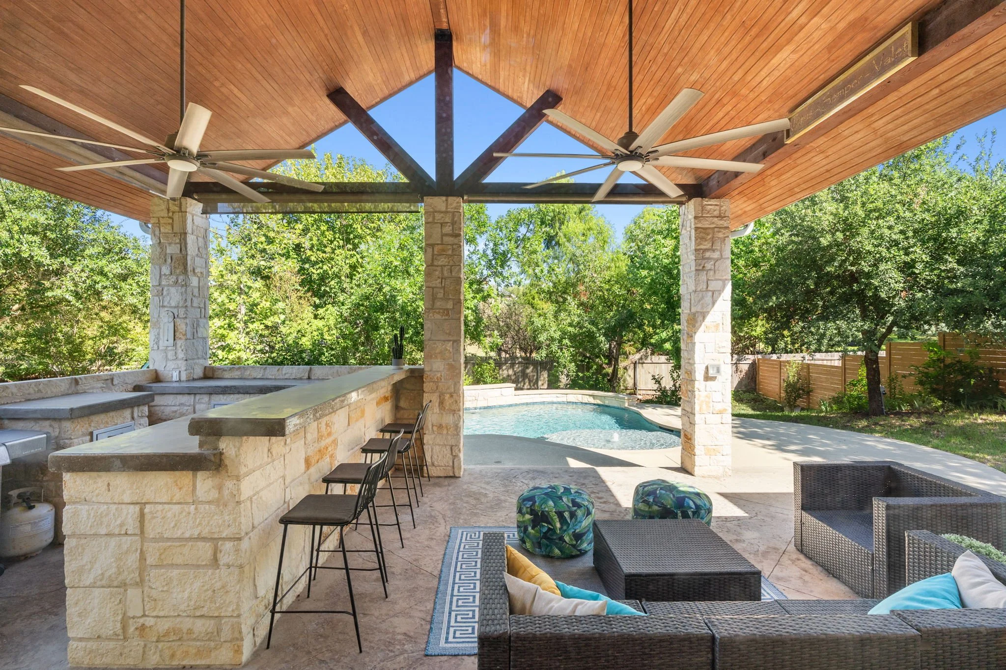Backyard patio with a stone countertop bar, chairs, a pool, surrounded by trees and a wooden fence, under a wooden roof with ceiling fans.