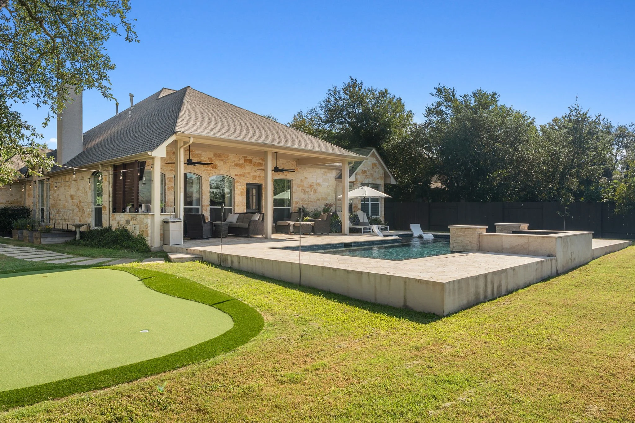 A backyard with a swimming pool, outdoor furniture, a patio, and a stone house with a covered porch under a clear blue sky.