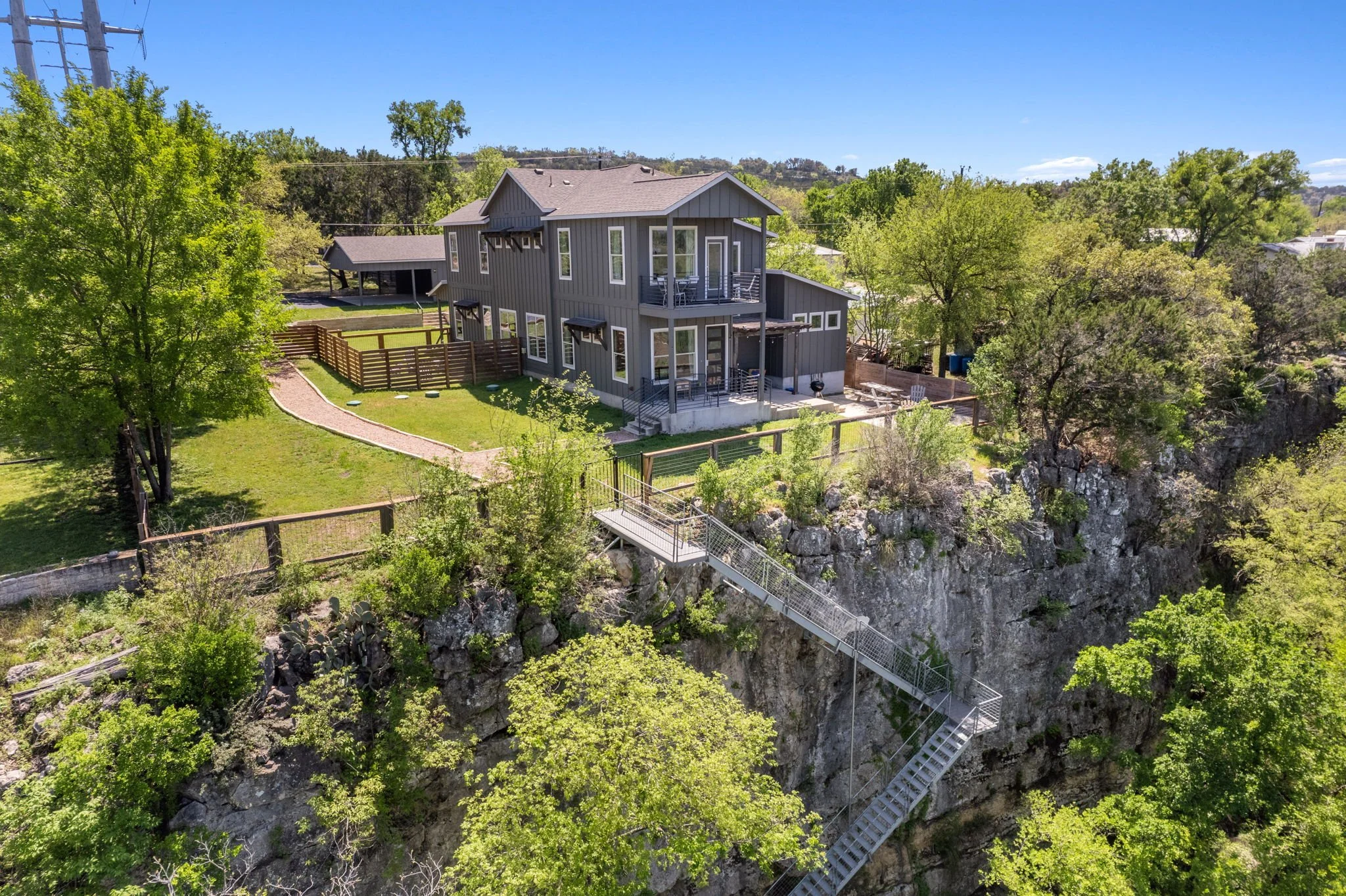 A modern gray house situated on a rocky hilltop with lush green trees surrounding it and a metal staircase descending from the house to the rocky ground below.