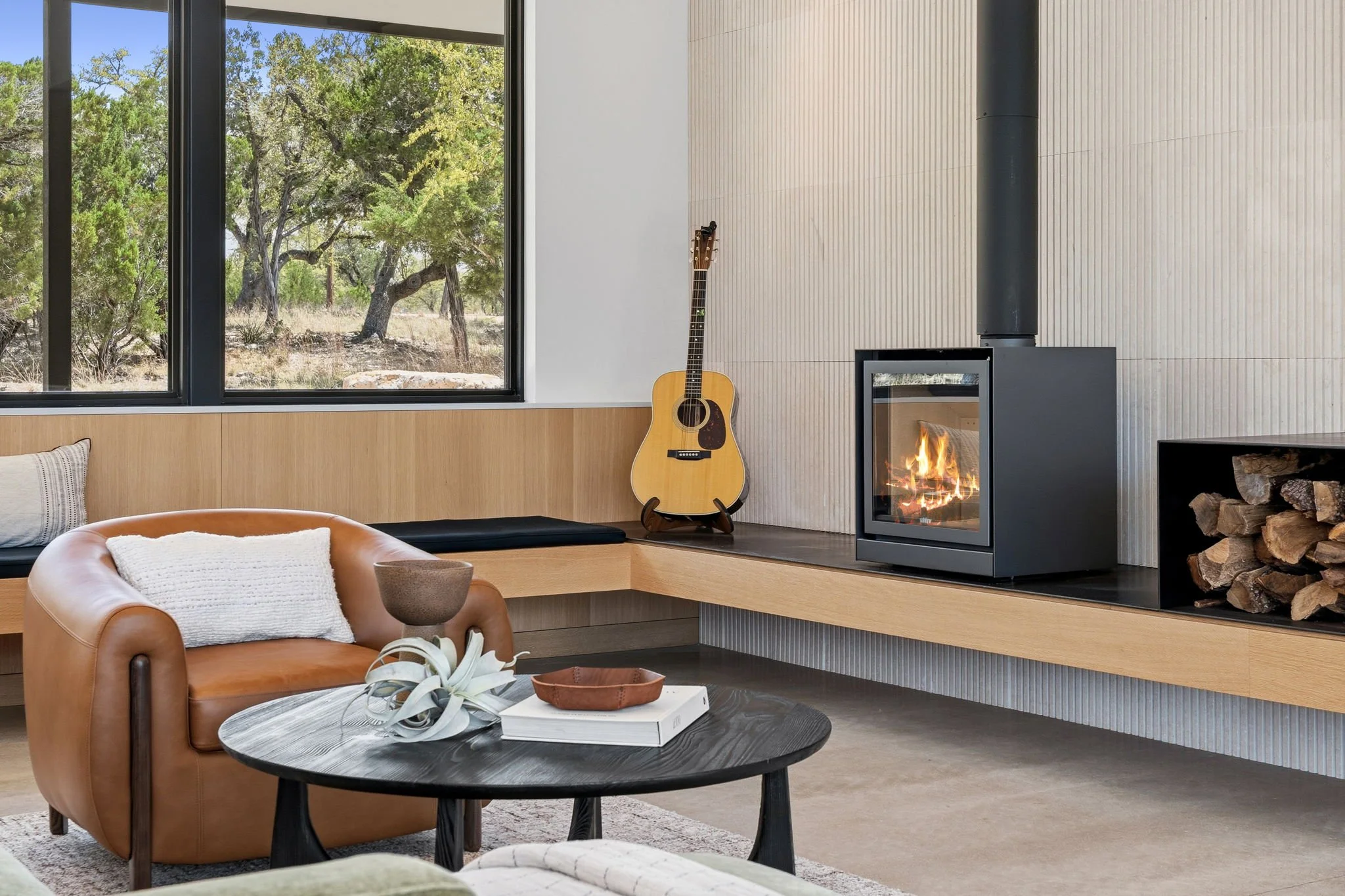 Modern living room with large window showing trees outside. The room has a cozy fireplace, a guitar leaning against the wall, and contemporary furniture including a leather armchair and a round coffee table with decor items.