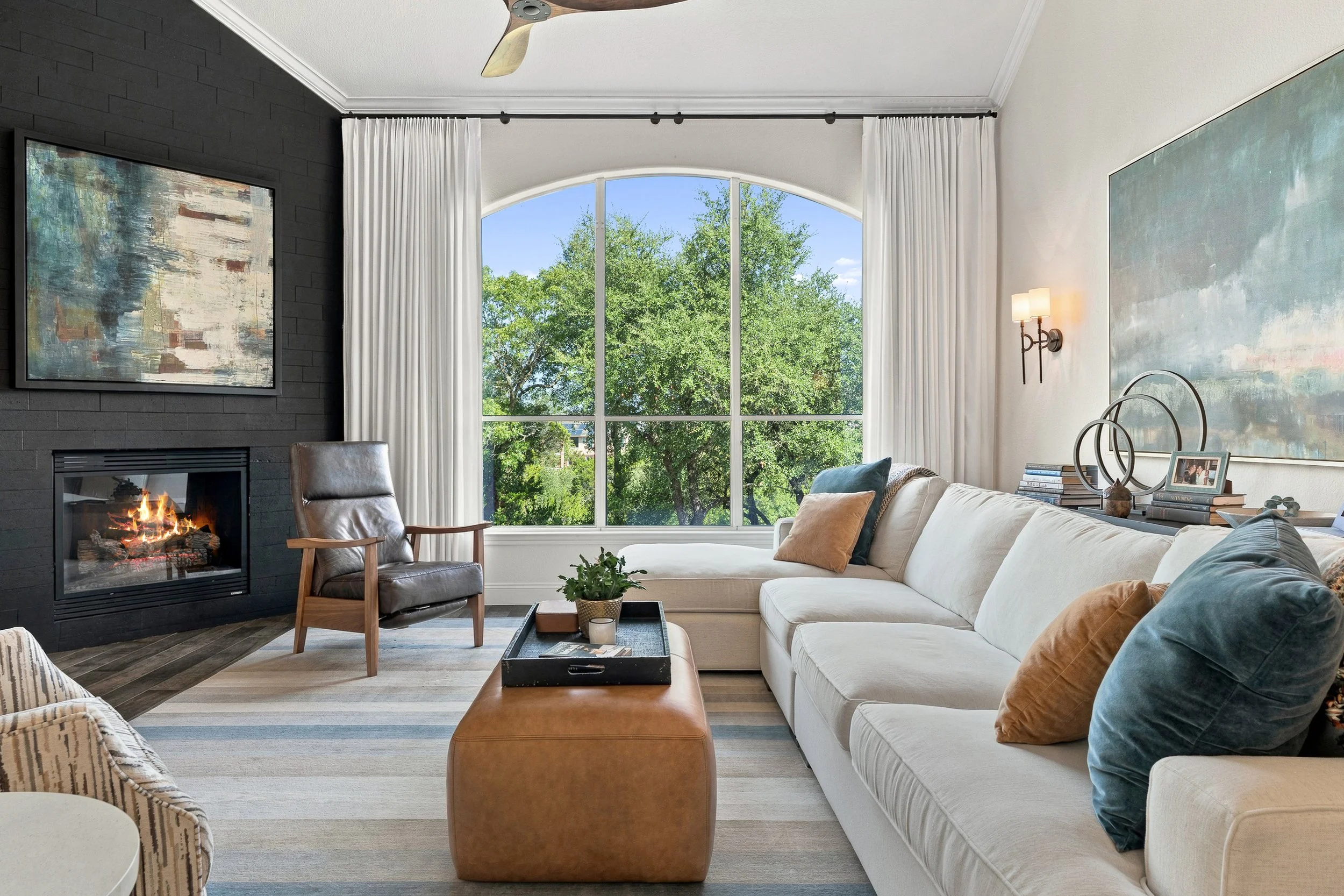 Living room with large window showing green trees, a white sofa with pillows, a coffee table with decor, a fireplace with fire, an armchair, and wall art.