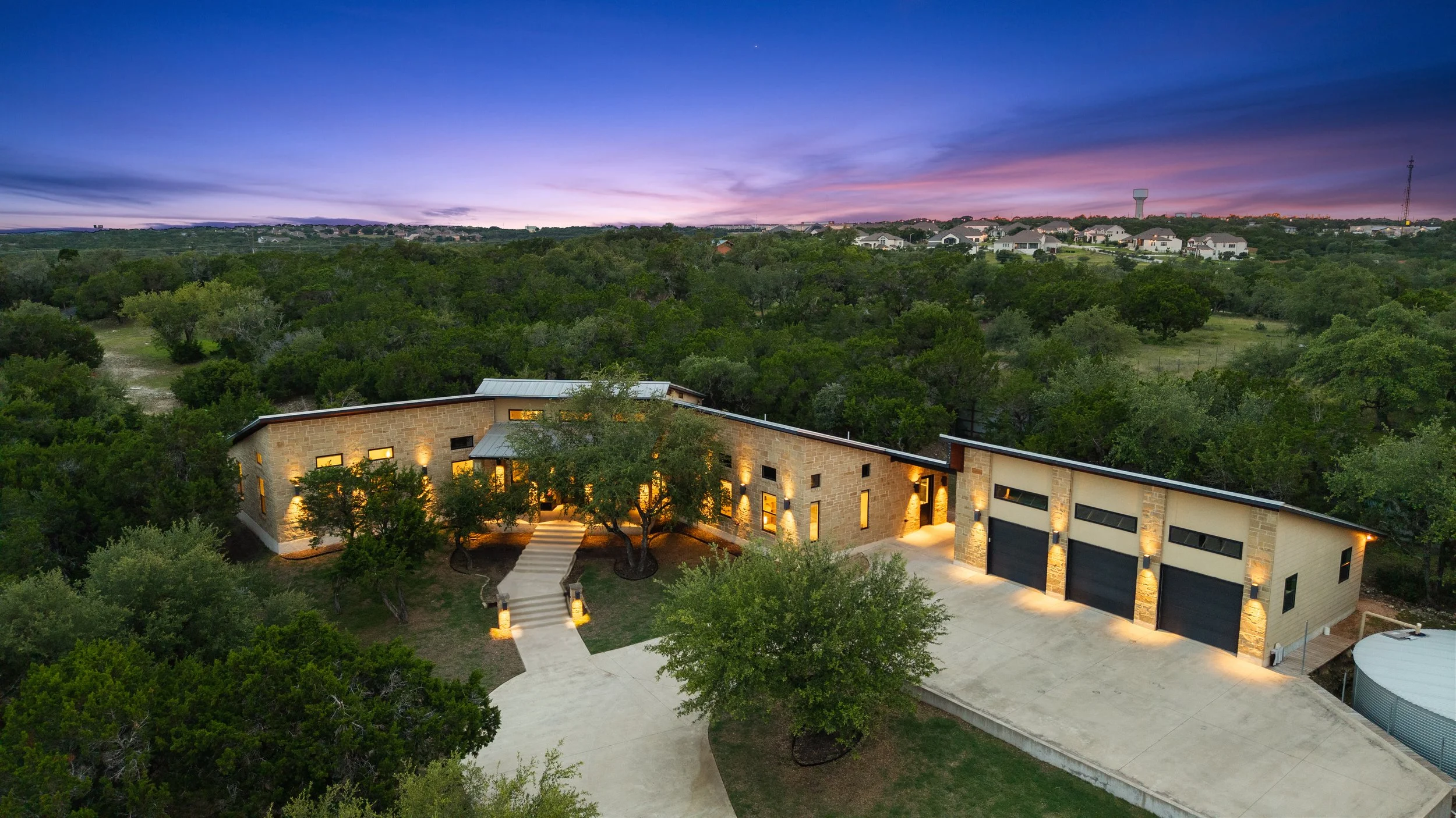 A modern house surrounded by greenery under a twilight sky with purple and blue hues, with the house's interior lights on and a driveway leading to three garage doors.