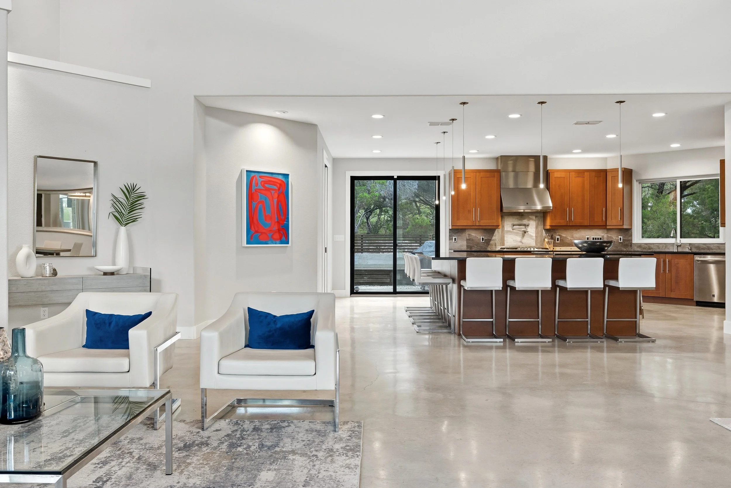 Modern open-concept kitchen and living space with white walls, wooden cabinets, stainless steel appliances, and large windows.
