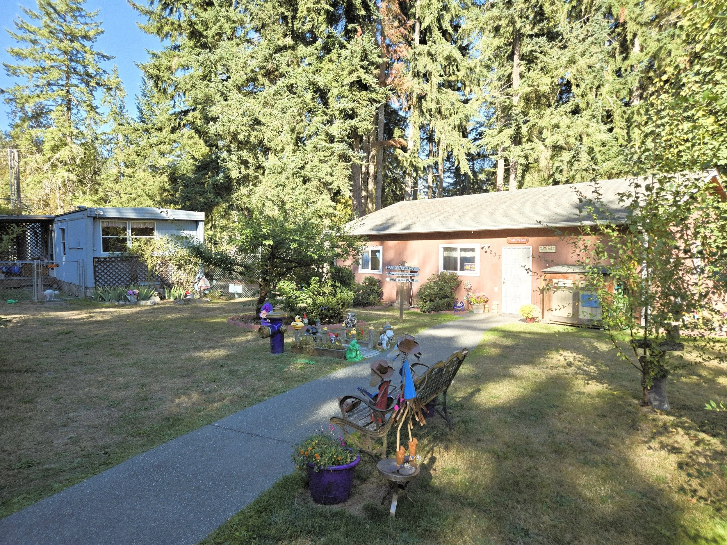 A shop with a light red exterior surrounded by lush green trees. A paved pathway leads to the front door. Decorated with signs, flowers, and garden ornaments, indicating it is pet-friendly and home of Paw Prince dog grooming and boarding facility.