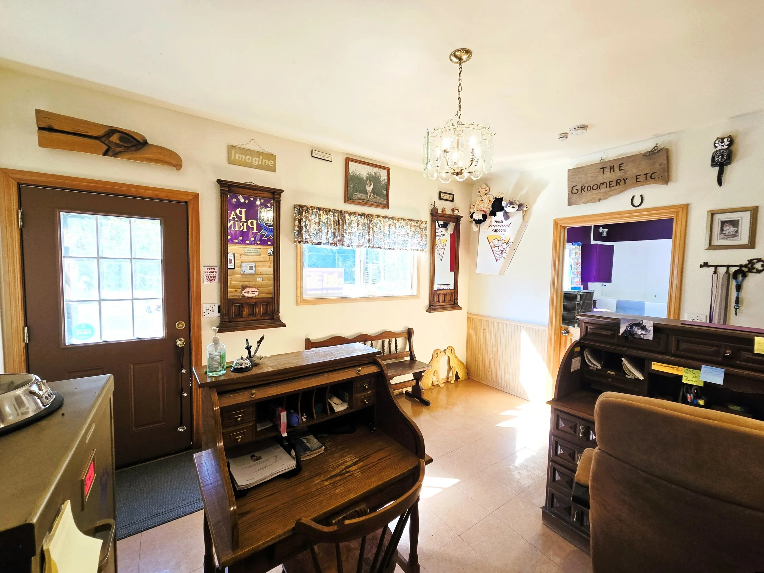 Interior of a cozy room with wooden furniture, a chandelier, and wall decorations including signs, framed pictures, and plush toys. There is a door with a window and sunlight coming through the window.