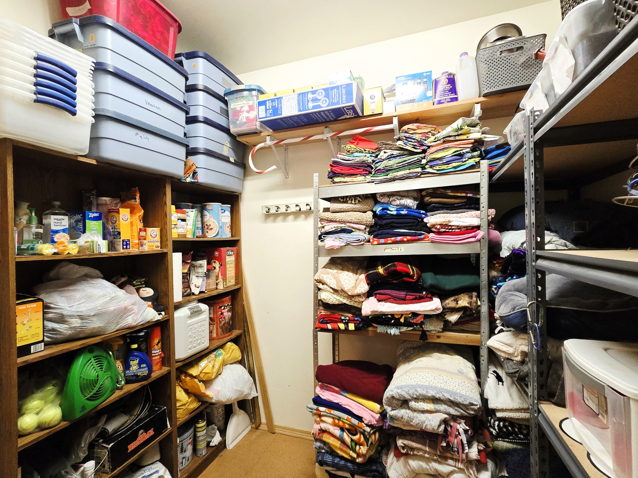 A storage closet with shelves filled with folded clothes, boxes, cleaning supplies, and household items.