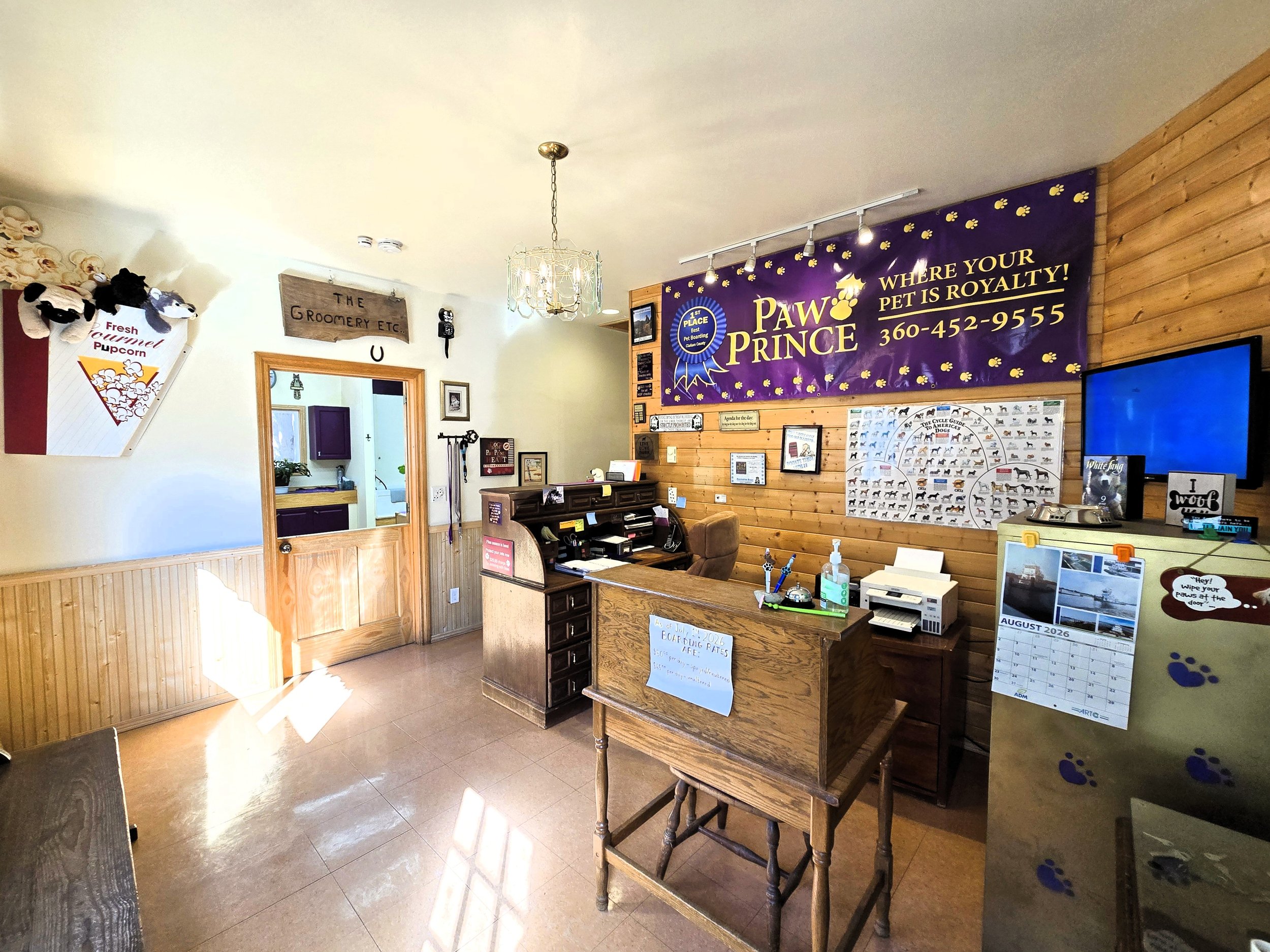 Indoor reception area of a pet grooming and boarding facility a purple banner that reads 'Paw Prince' and a phone number, wooden walls, a reception desk, and various pet-related displays and signs.