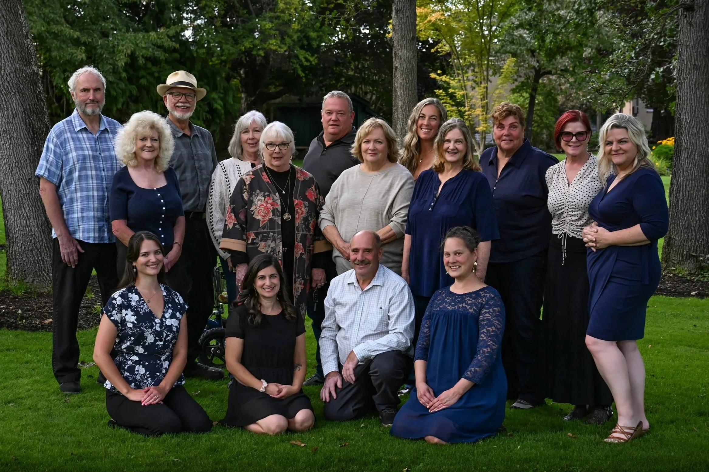 A group of real estate professionals posing outdoors for a photo among trees and green grass. Smiling and ready to serve.