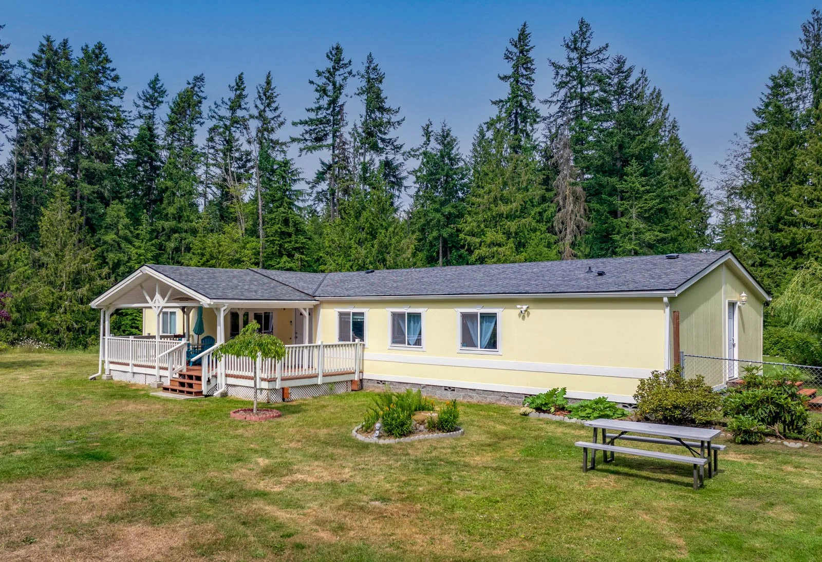 A single-story yellow house with a dark shingle roof, surrounded by green trees and a well-maintained lawn. The house features a front porch with white railings and a small set of stairs, and there is a picnic table in the yard.