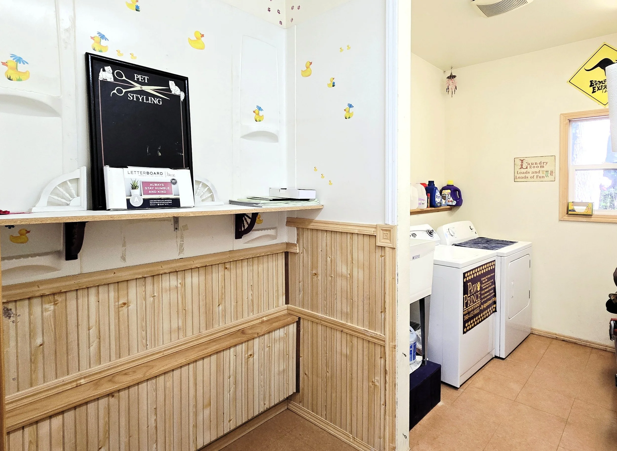 A laundry room with a washer and dryer, a wooden wainscoting, cleaning supplies on a shelf, and a window. There are decorative duck stickers on the wall and signs related to pet grooming and a dog training school.