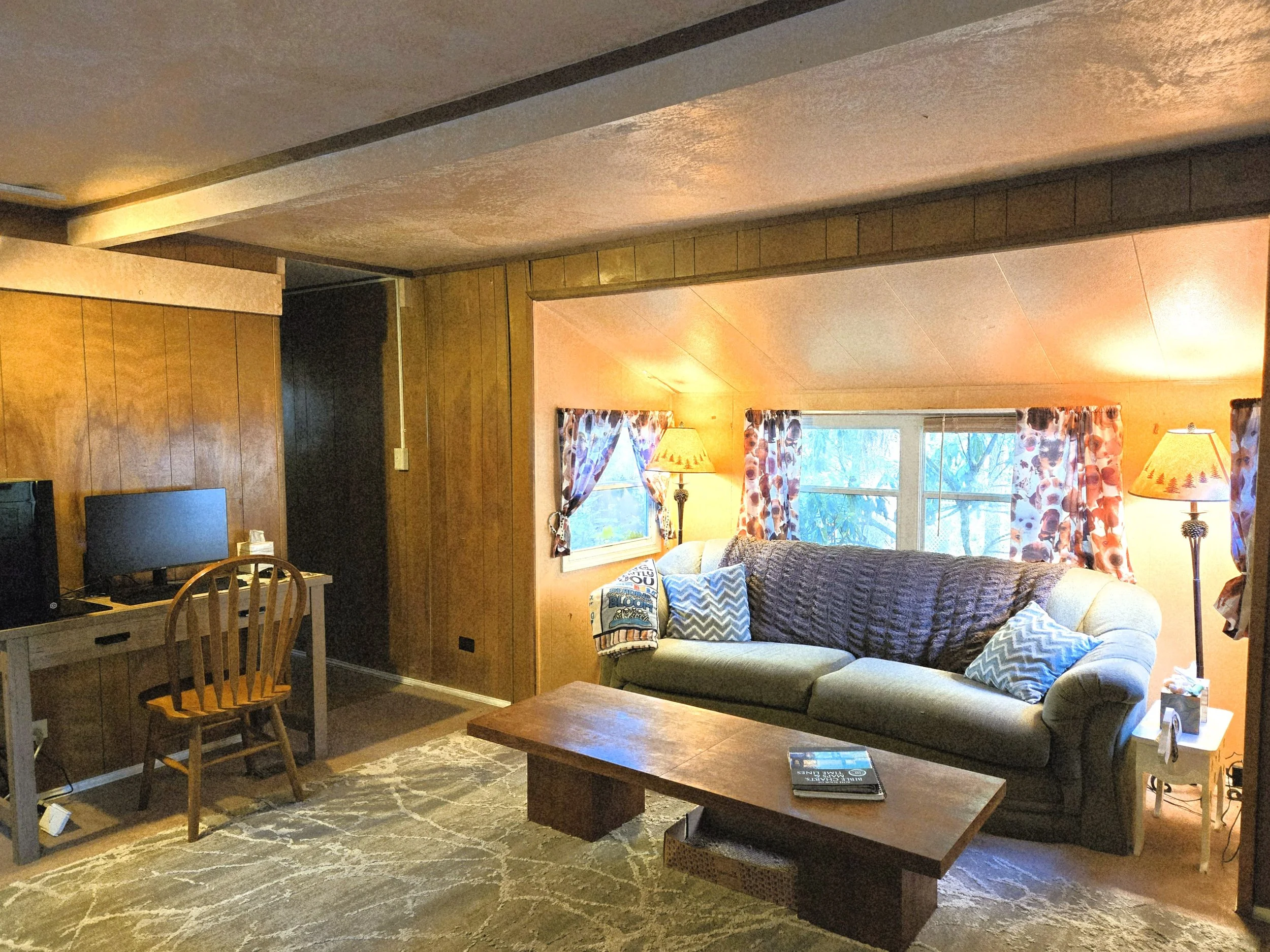Living room with wooden walls, a beige sofa with pillows, a wooden coffee table, a window with floral curtains, two lamps, and a television on a wooden desk.