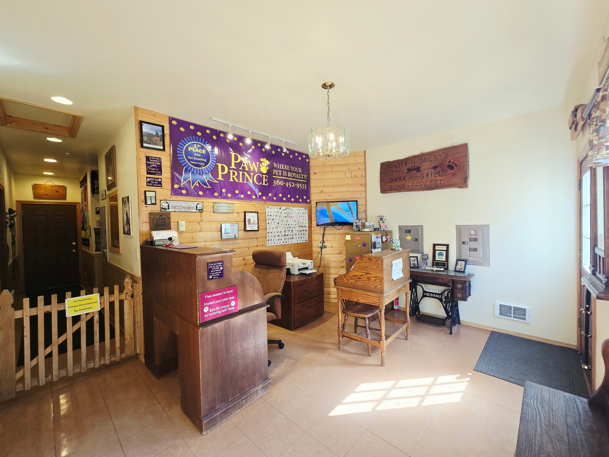 Reception area of a pet boarding facility with signs advertising pet services, a wooden desk, chairs, and a small fenced area.