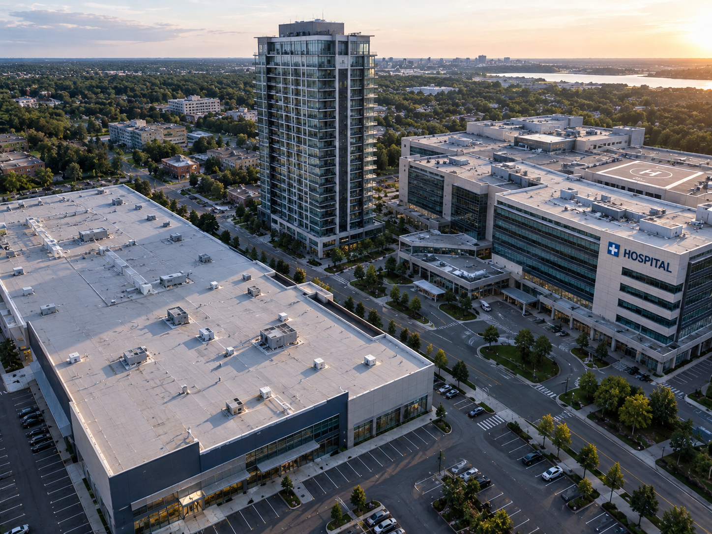 Aerial view of a hospital complex with surrounding greenery and cityscape in the background during sunset. The hospital has multiple stories with a heliport on the roof, and adjacent buildings include a tall glass tower and a large flat-roofed structure.