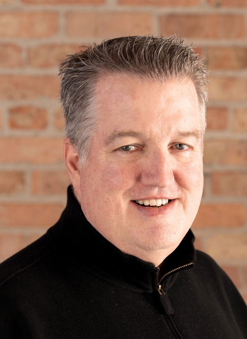 Portrait of a smiling middle-aged man with short, grayish hair, wearing a black zip-up sweater, standing in front of a brick wall.