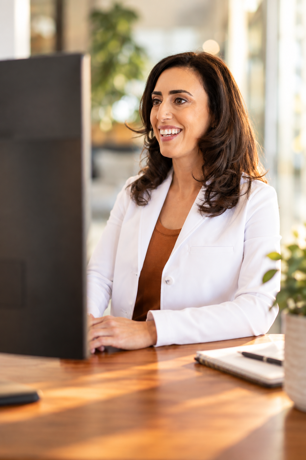 Alani Kalfayan in a white blazer working on a desktop computer at a wooden desk in a bright office.