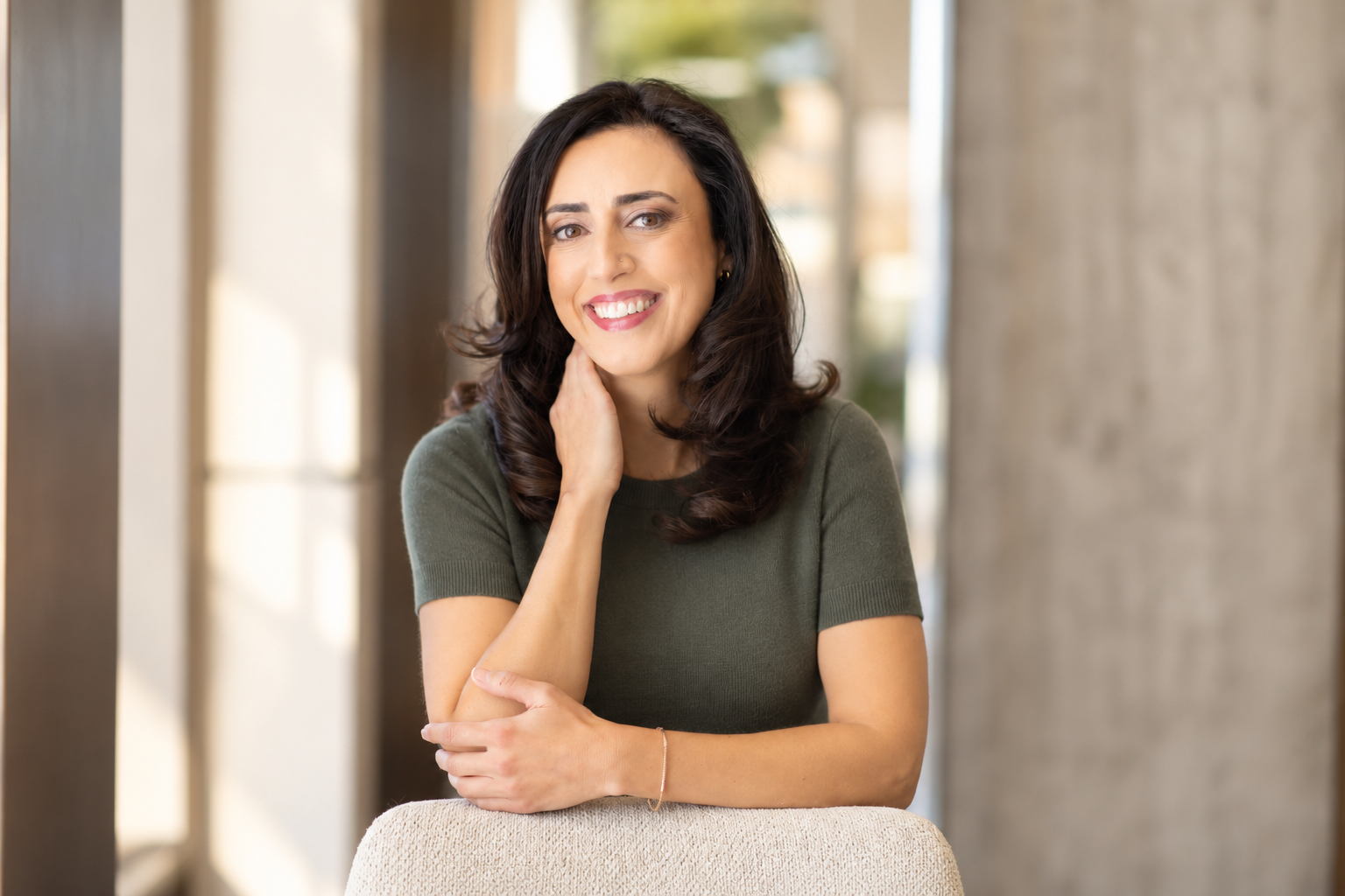Alani Kalfayan with dark brown hair, smiling, wearing a green short-sleeve top, sitting indoors with blurred background.