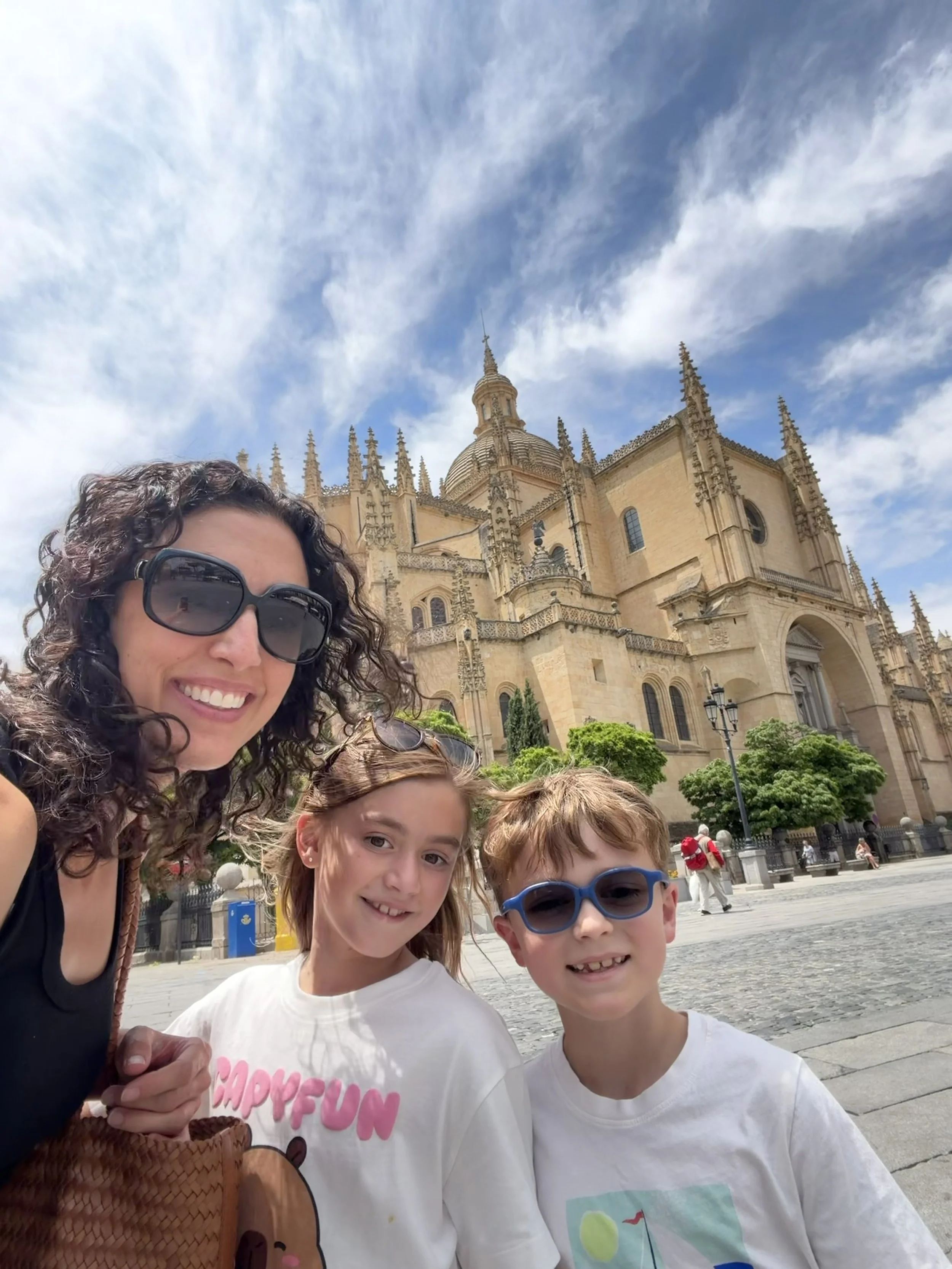 A woman with curly hair and sunglasses smiling with three children, standing in front of a historic building with a cloudy sky above.