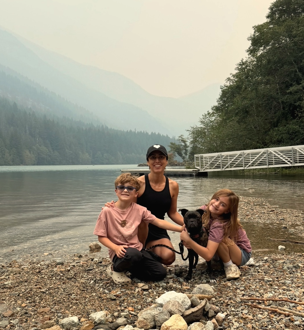 A woman and two children with a dog by a lake with mountains and trees in the background.