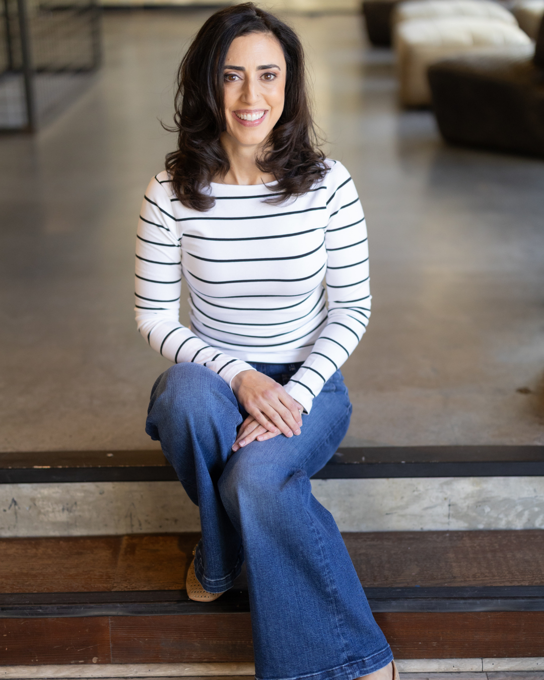 Alani Kalfayan with dark wavy hair sitting on a step, smiling, wearing a white and black striped long sleeve shirt and blue jeans, in a cozy indoor setting.