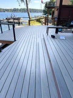 Long runs of gray decking form the main surface beside the home, with contrasting boards outlining the edges. This stage highlights the board layout and transitions around the deck opening as installation continues.