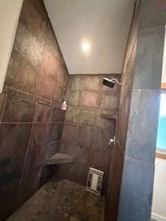 This shower remodel combines dark wall tile with a built-in corner bench and decorative accent details. The tiled surfaces establish the bathroom’s richer finish as work continues toward completion.