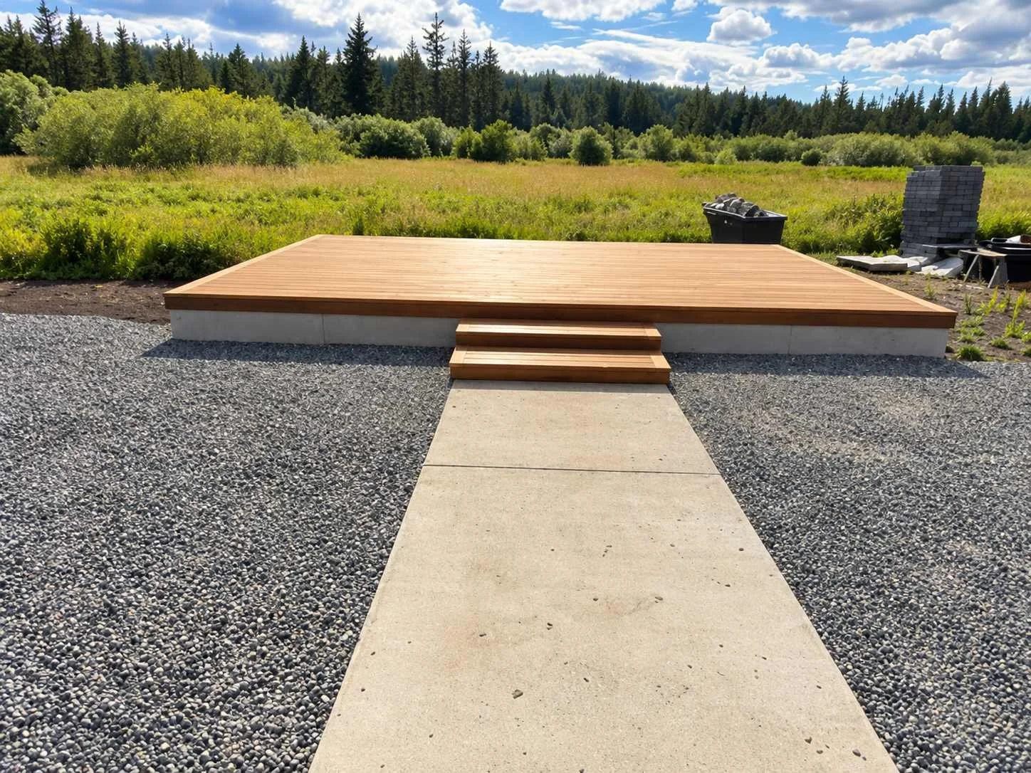A new freestanding wood deck creates a gathering space connected to the existing concrete walkway. Broad entry steps provide a transition onto the platform, with gravel surrounding the deck.