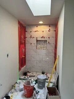The shower renovation is underway with wall tile being installed around a recessed storage niche. Exposed wall preparation and installation materials show the project before the remaining tile and fixtures are completed.
