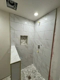 Light marble-look tile wraps the shower walls, complemented by a recessed niche and a smaller mosaic floor pattern. This stage brings the main tile finishes together before the remaining fixtures and final details are installed.