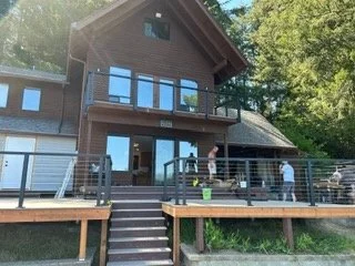The deck extends across the waterfront side of the home, with a central staircase connecting it to the ground below. Decking and railing installation bring the main outdoor living area together as work continues.