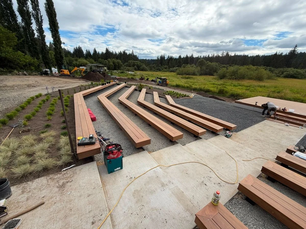 This outdoor gathering-space project includes multiple rows of angled wood benches and a raised platform with steps. Construction is underway alongside surrounding landscaping and gravel areas.