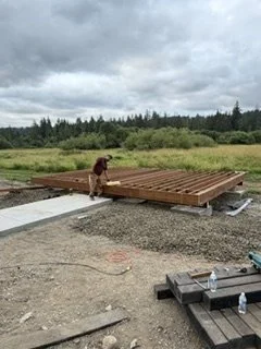 The freestanding deck begins with a wood framework beside the existing concrete walkway. The crew is assembling the platform’s supports and joists before adding deck boards and the front entry steps.