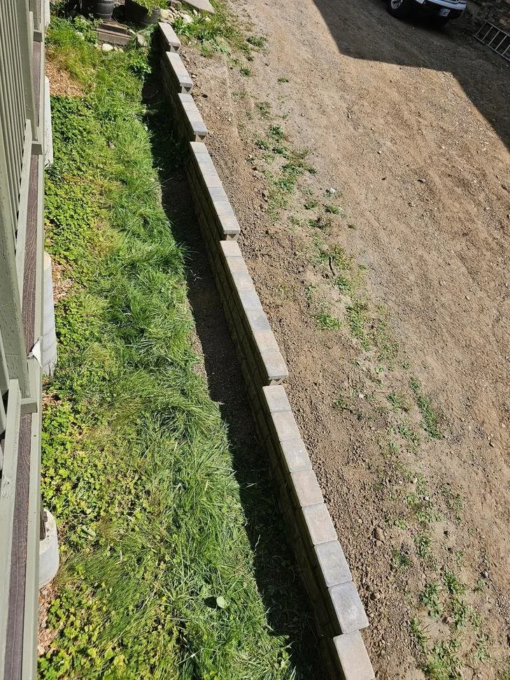 A straight block retaining wall separates the raised yard from the narrow strip alongside the home. The installation follows the building’s side, creating a defined edge between areas at different elevations.