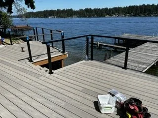 Deck boards and black railings are being installed around the waterfront platform and stair opening. The open railing design keeps the water visible while defining the edges of the outdoor space.