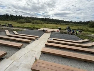 The gathering-space layout places wood benches on both sides of a central concrete walkway. The path connects the seating area to the platform while dividing the bench sections.