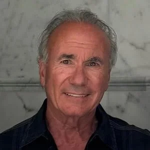A headshot of Nick Vlachos with gray hair, smiling, wearing a dark shirt, against a gray blurred background.