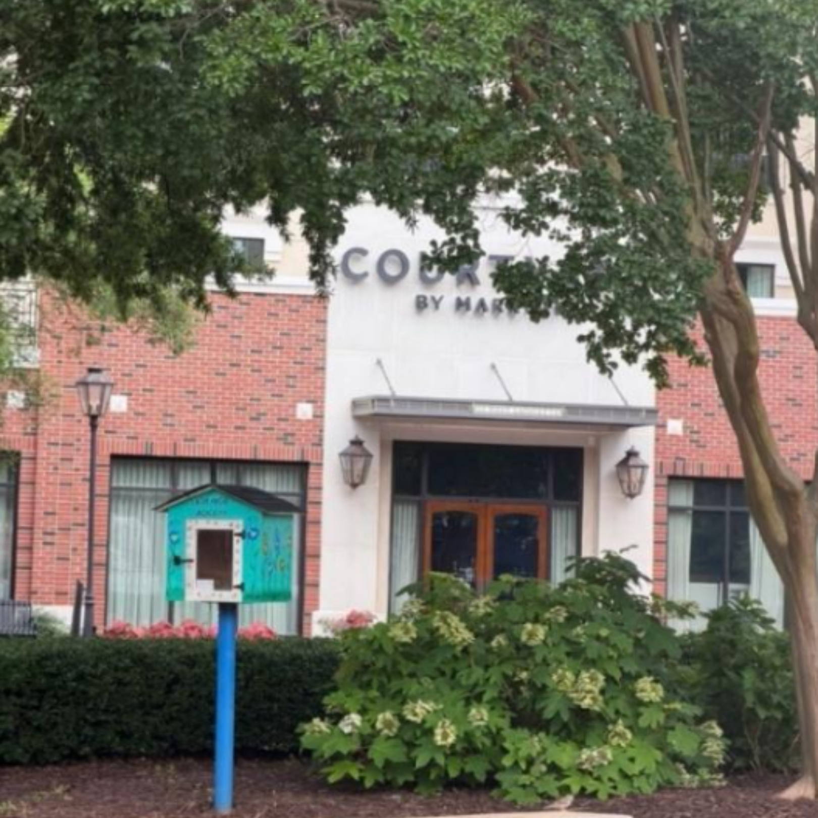 View of a building labeled 'COUTURE BY MAR' partially obscured by tree branches in front. There is a small blue structure that looks like a kiosk or booth with a sign on it, and decorative flowers and bushes at the base of the building.