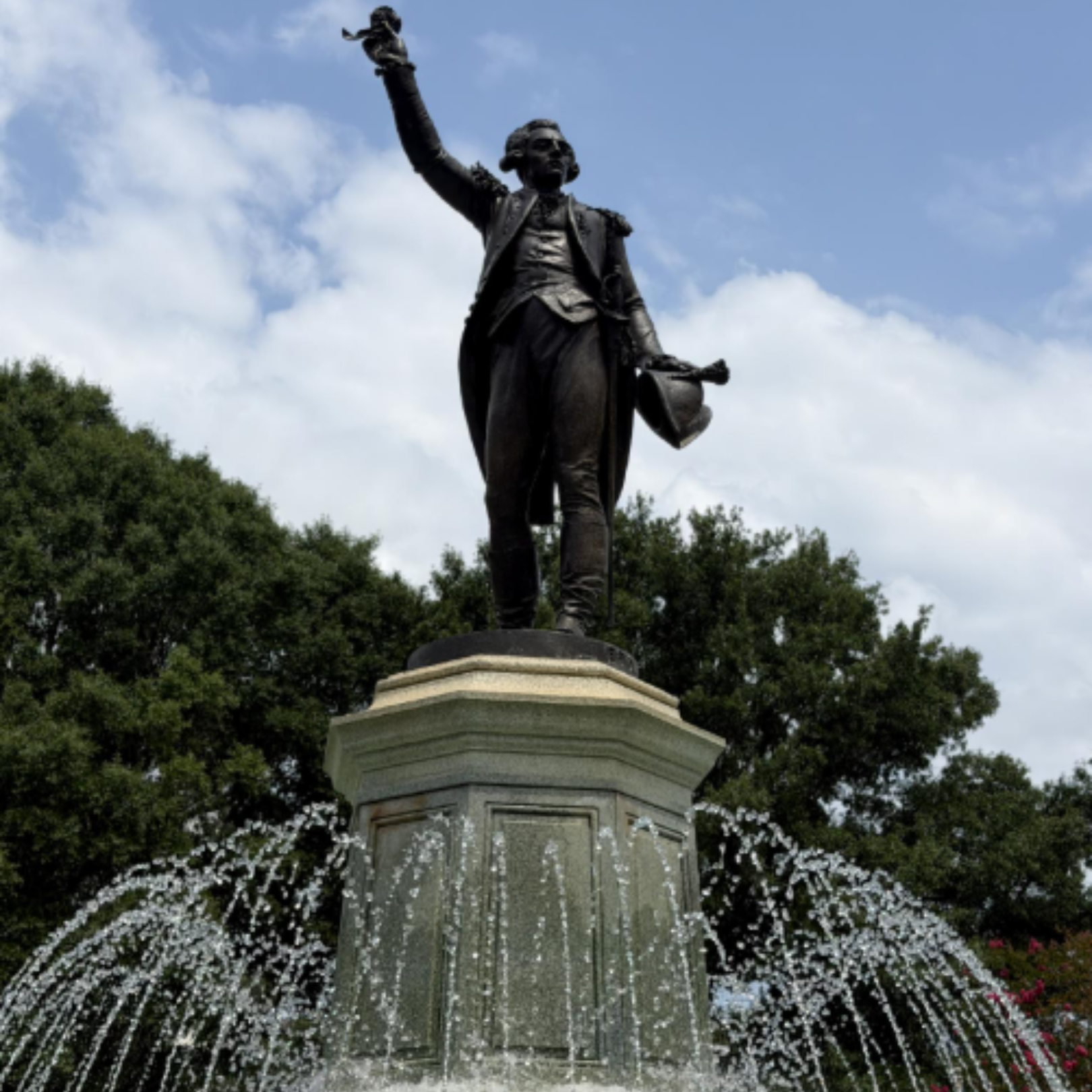 Statue of a historical figure on a pedestal, surrounded by a fountain with water spraying outward, greenery and trees in the background, under a partly cloudy sky.