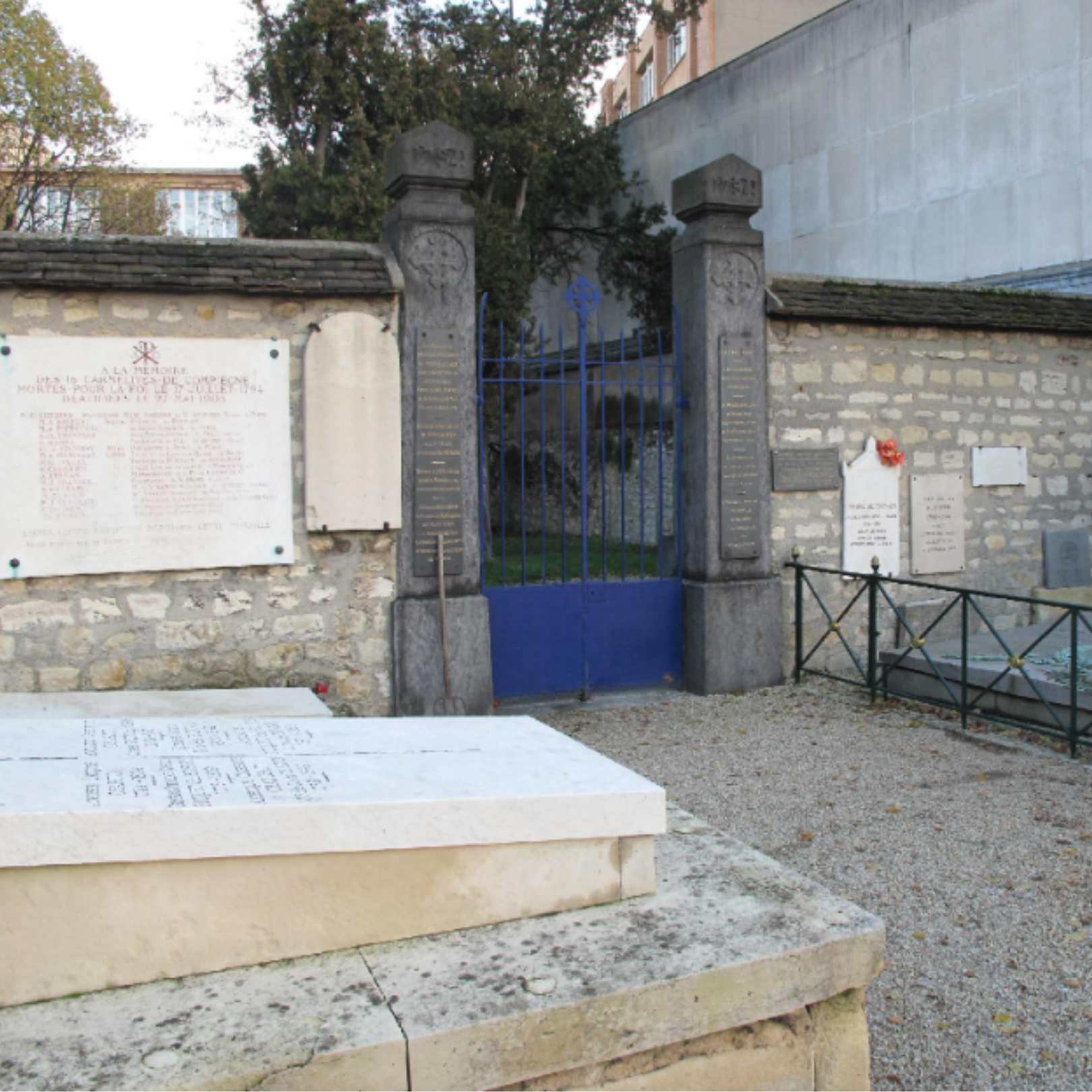 Gravestone and memorial plaques at a historic site, with a metal gate and stone wall, framed by trees and buildings in the background.