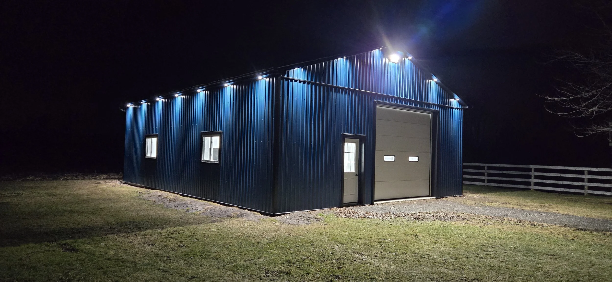 A blue metal building with a garage door and a side door, illuminated by outdoor lights at night, with a grassy area and a wooden fence nearby.