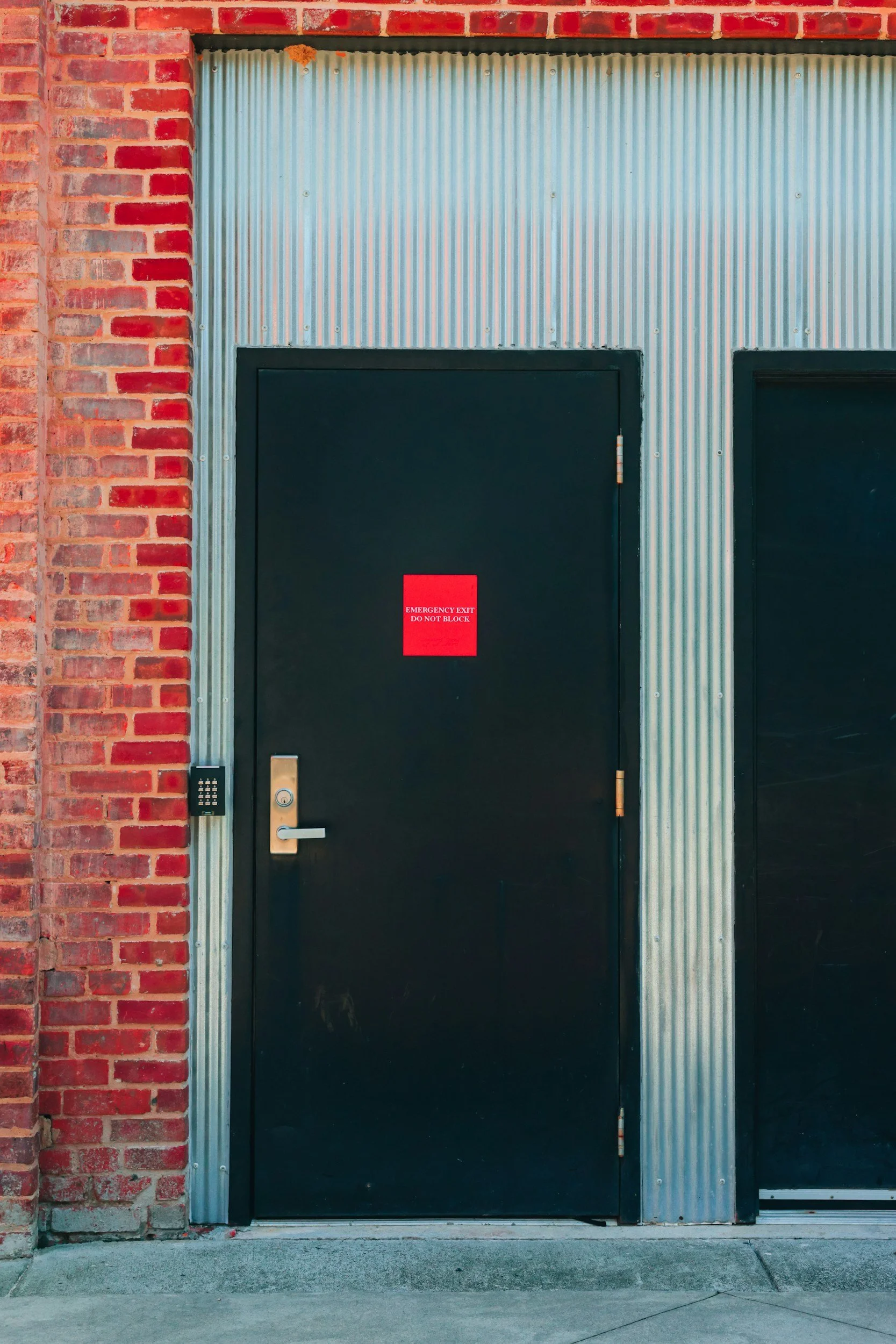 Black emergency exit door with a red sign that reads 'Emergency Exit Do Not Block', set in a building with a red brick wall on the left and corrugated metal framing around the door.