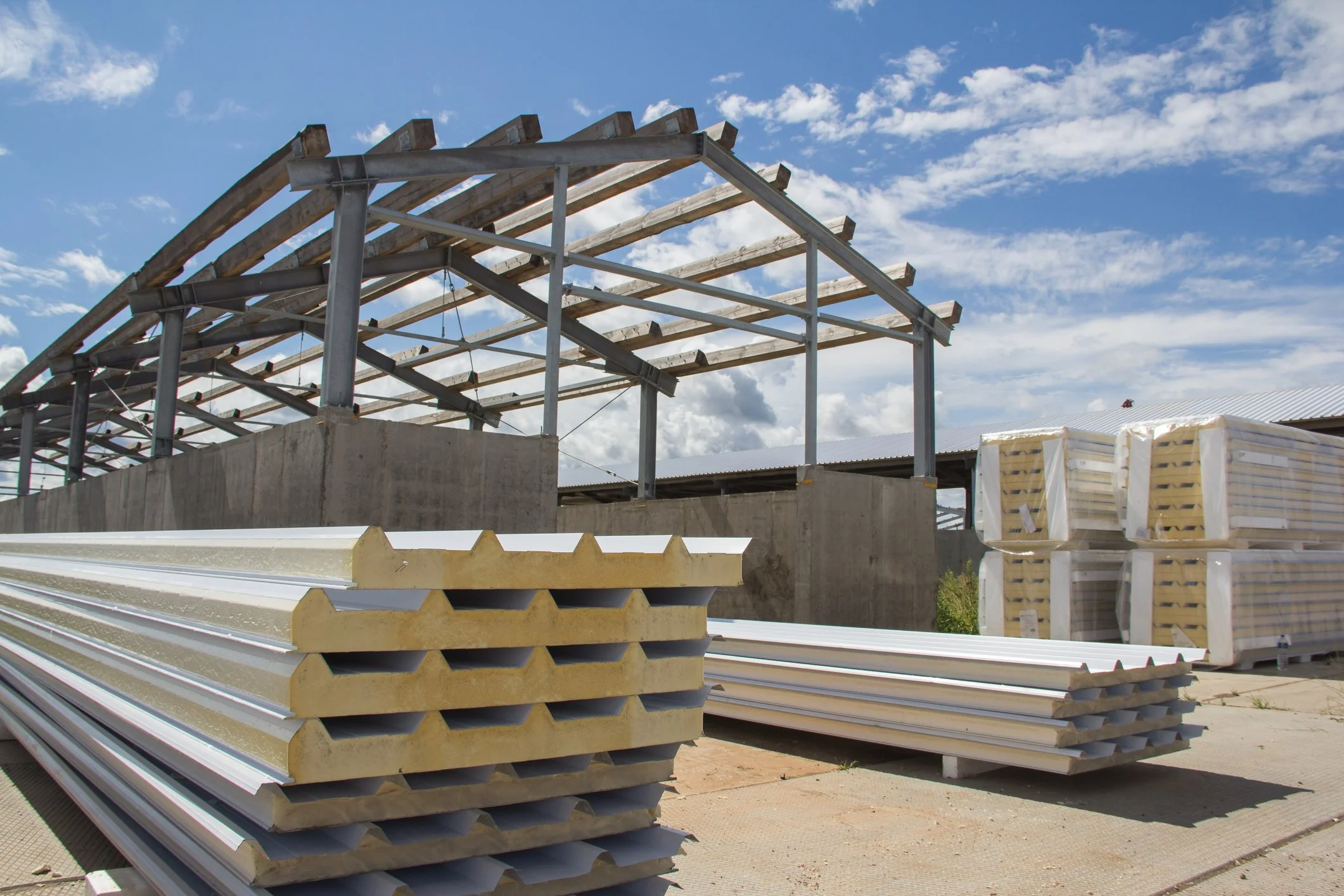 Stacks of construction materials, including metal and insulated panels, at a building site with a partly built structure and a blue sky with clouds in the background.