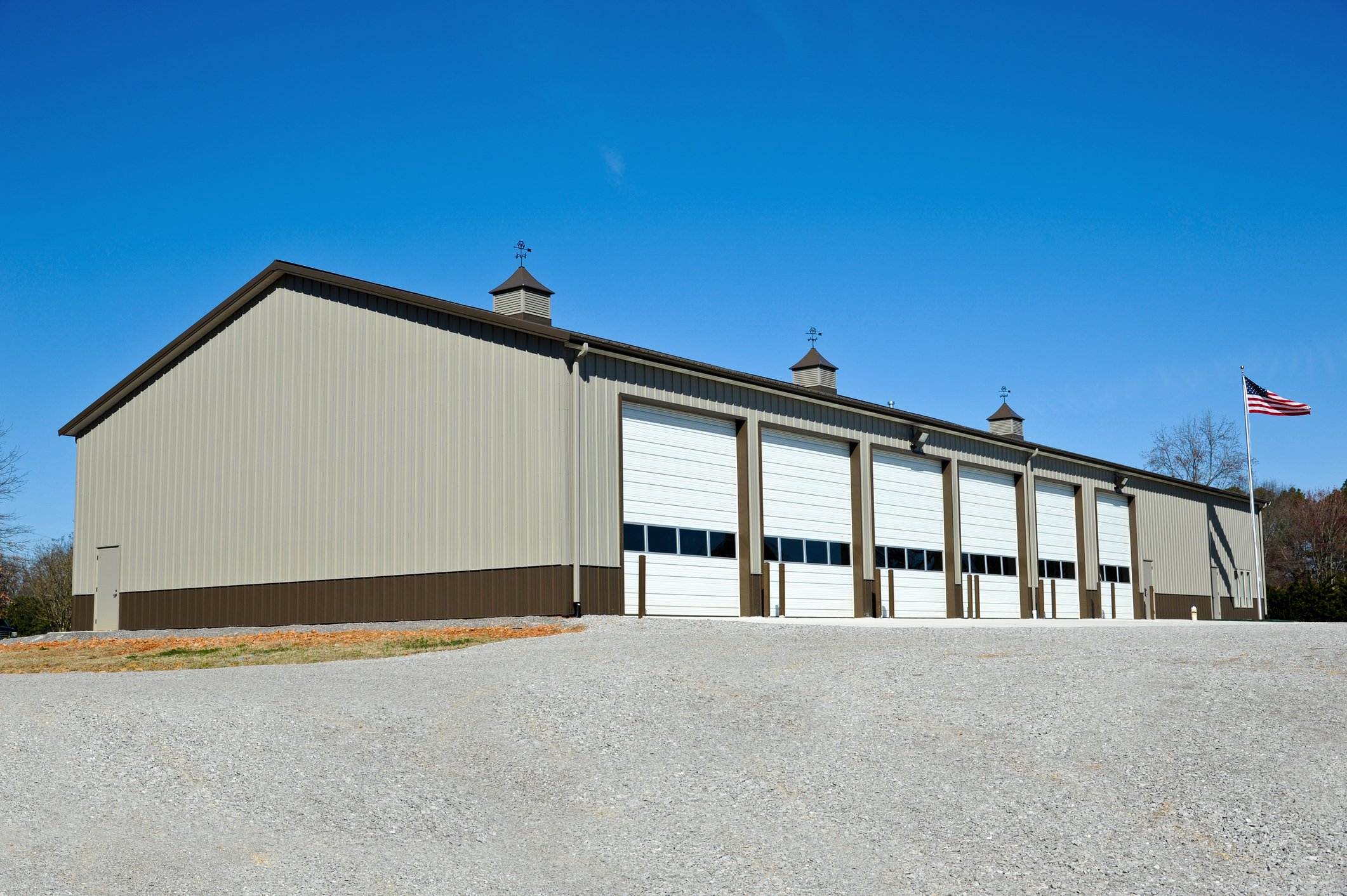 Large metal storage building with multiple garage doors, beige siding, and three small cupolas on the roof, with an American flag flying nearby against a blue sky.
