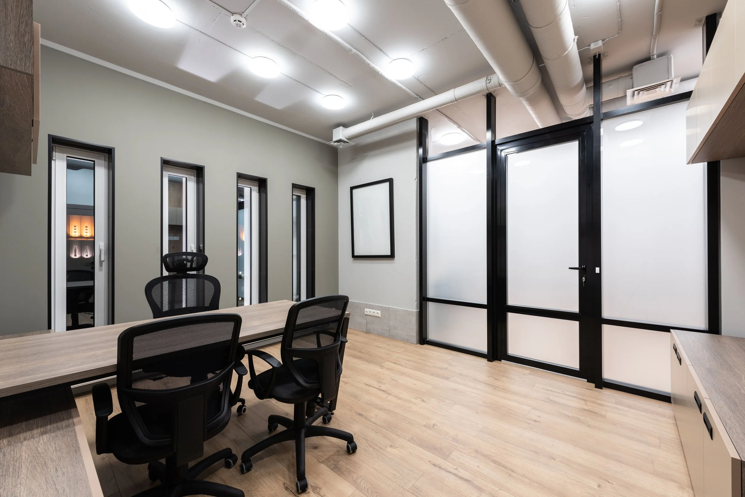 A modern office conference room with a wooden table, black mesh chairs, a white wall, and multiple windows with frosted glass. The ceiling has exposed pipes and circular ceiling lights.
