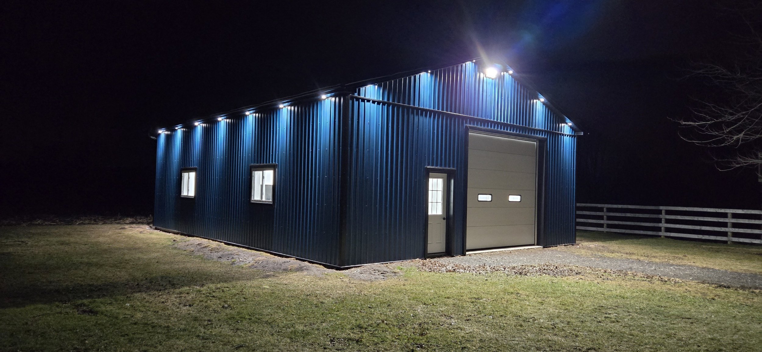A dark blue metal barn illuminated by exterior lights at night, with a large garage door, a side door, and three small windows, surrounded by a grassy area and a wooden fence in the background.