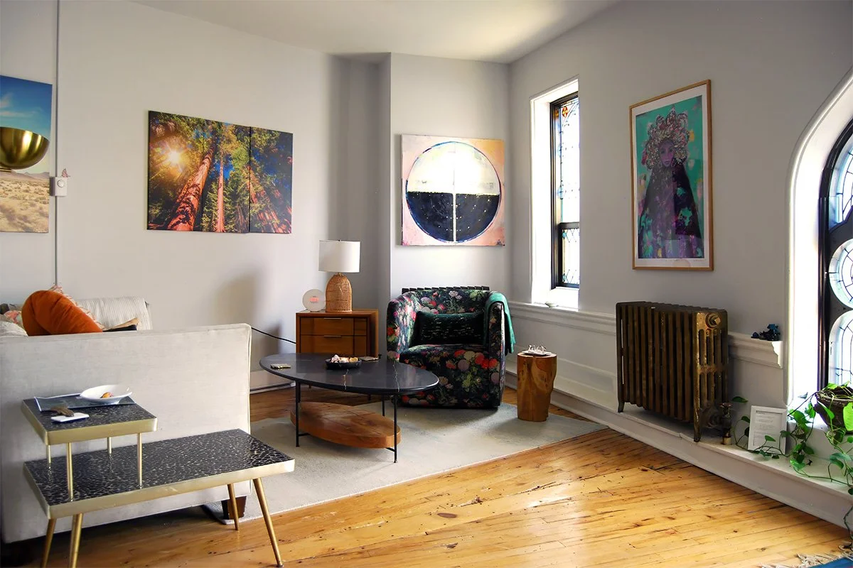 Bright eclectic therapist's office with colorful artwork, floral armchair, white sofa, coffee table, stained-glass windows, and wood floors