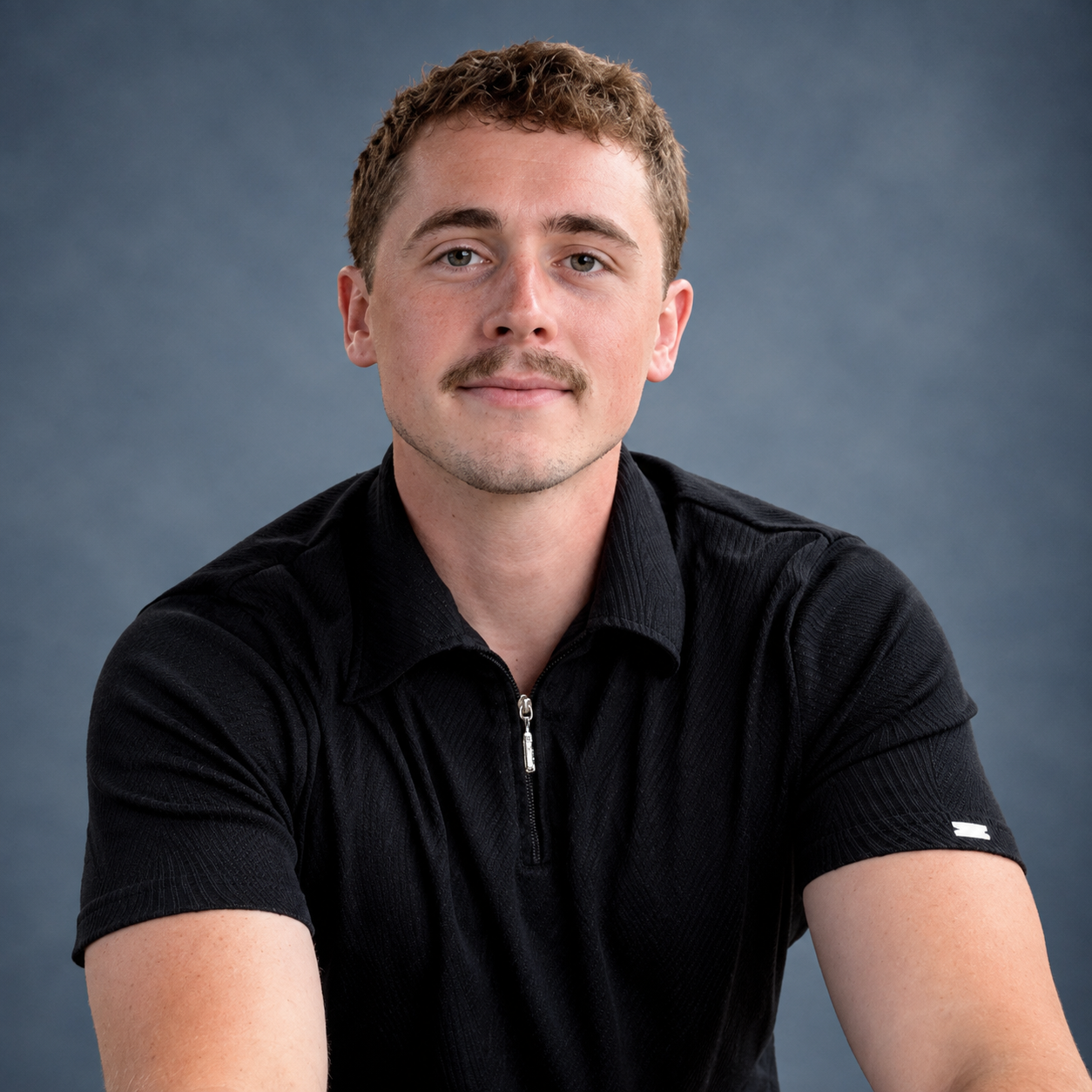 Portrait of a young man with short curly brown hair, light facial hair, wearing a black zip-up shirt, smiling slightly, against a gray background.