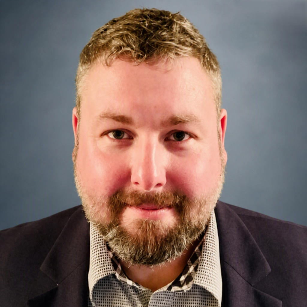 Close-up portrait of a man with short curly hair, a beard, and mustache, wearing a dark blazer over a patterned shirt, against a plain background.