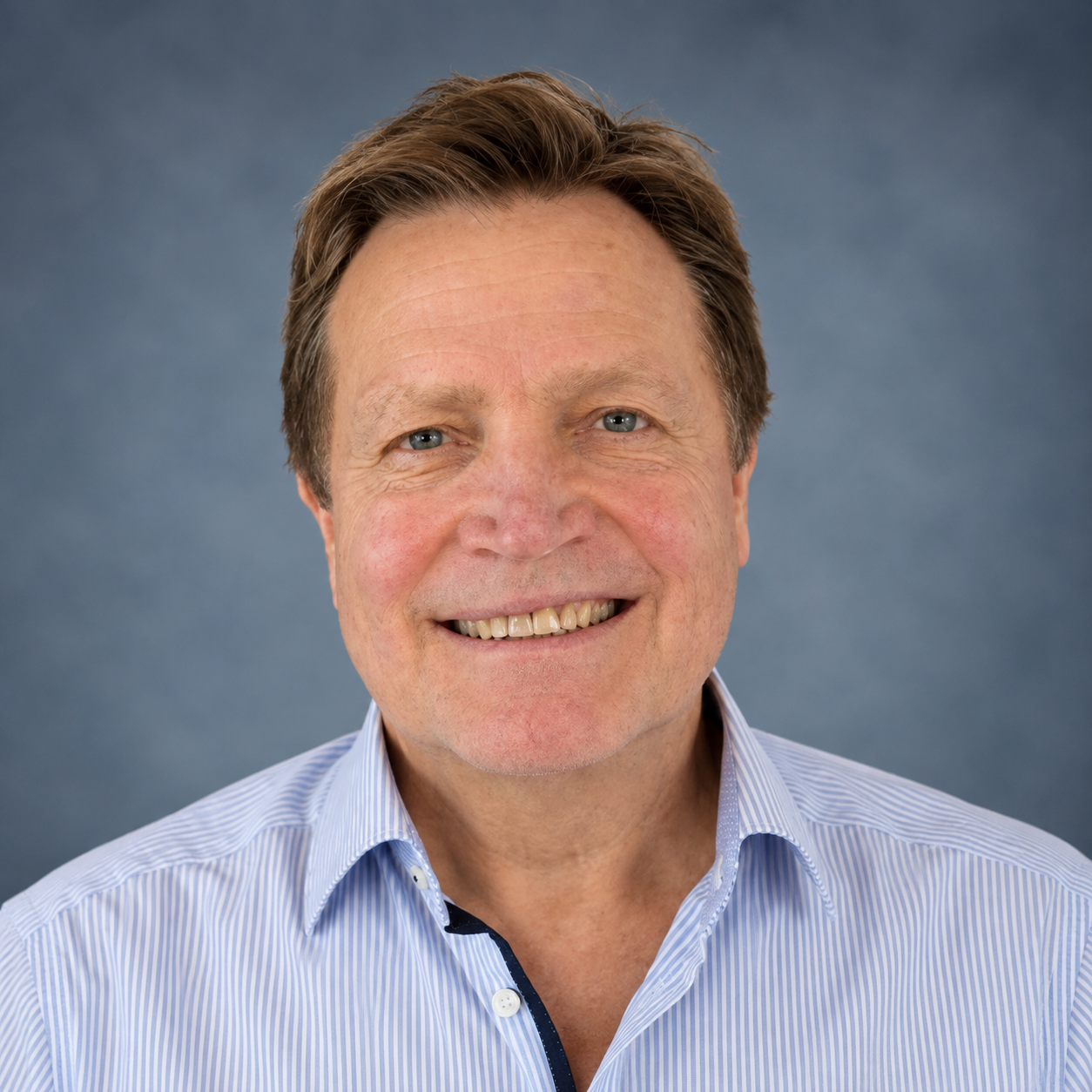 A smiling middle-aged man with light skin, short brown hair, wearing a blue and white striped collared shirt against a plain blue background.
